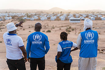 Vier UNHCR-Helfer*innen von hinten, auf Landschaft mit Zelten schauend