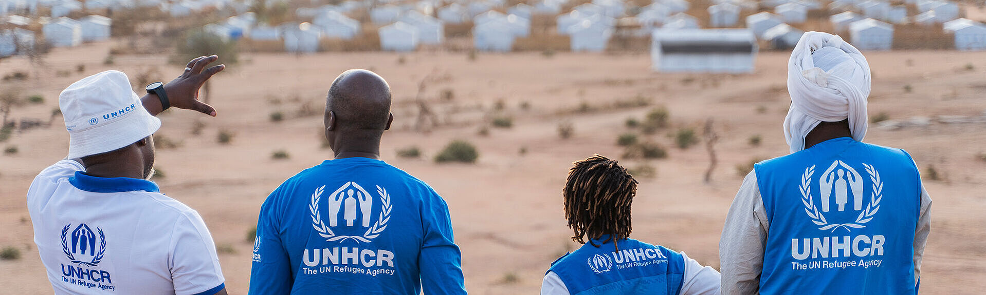 Vier UNHCR-Mitarbeiter*innen von hinten, schauen auf eine Landschaft mit Zelten.