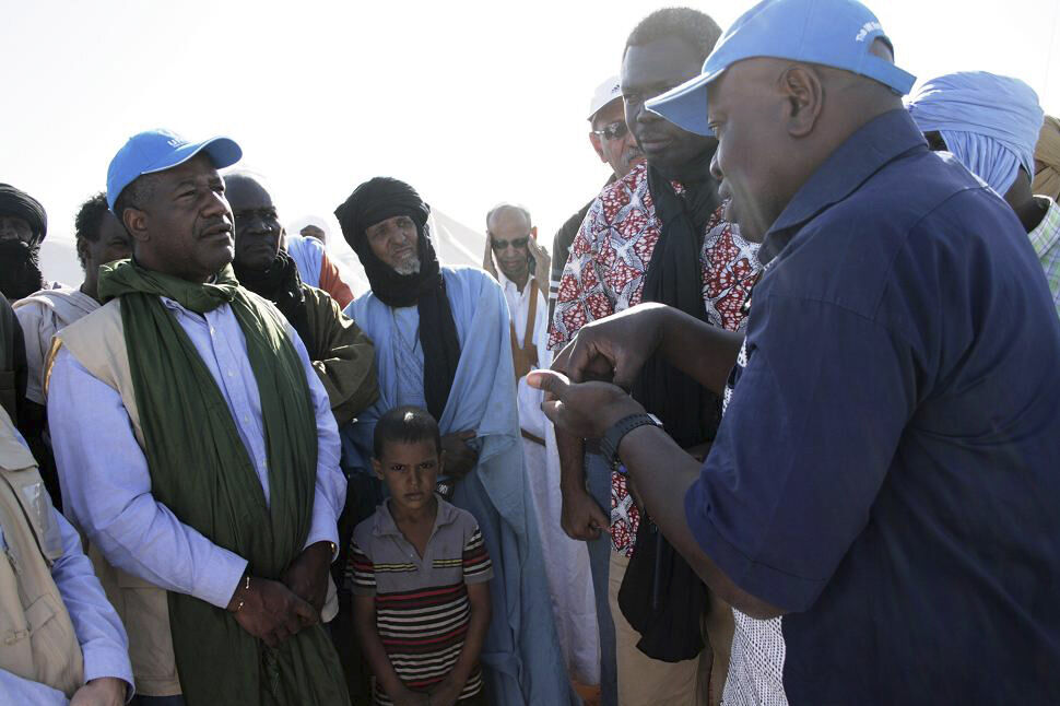 Valentin Tapsoba im Gespräch in einem Flüchtlingslager in Mauretanien. Man mit UNHCR Kappe spricht mit Flüchtlingen aus Mali. Zusehen ist eine Gruppe von Menschen.  