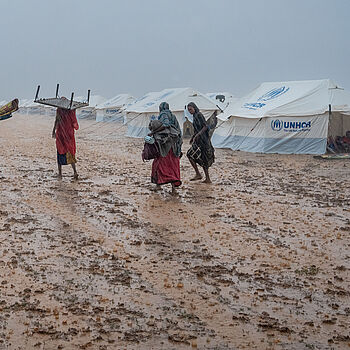 Frauen im Unwetter mit Gepäck und UNHCR Zelten im Hintergrund