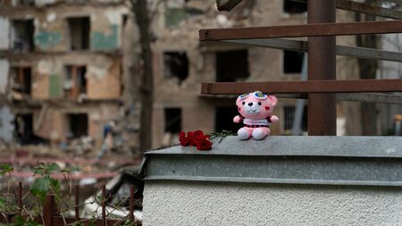 Ein Teddybär mit roten Rosen auf einem Gemäuer vor zerstörten Häusern 