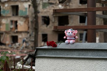 Ein Teddybär mit roten Rosen auf einem Gemäuer vor zerstörten Häusern 