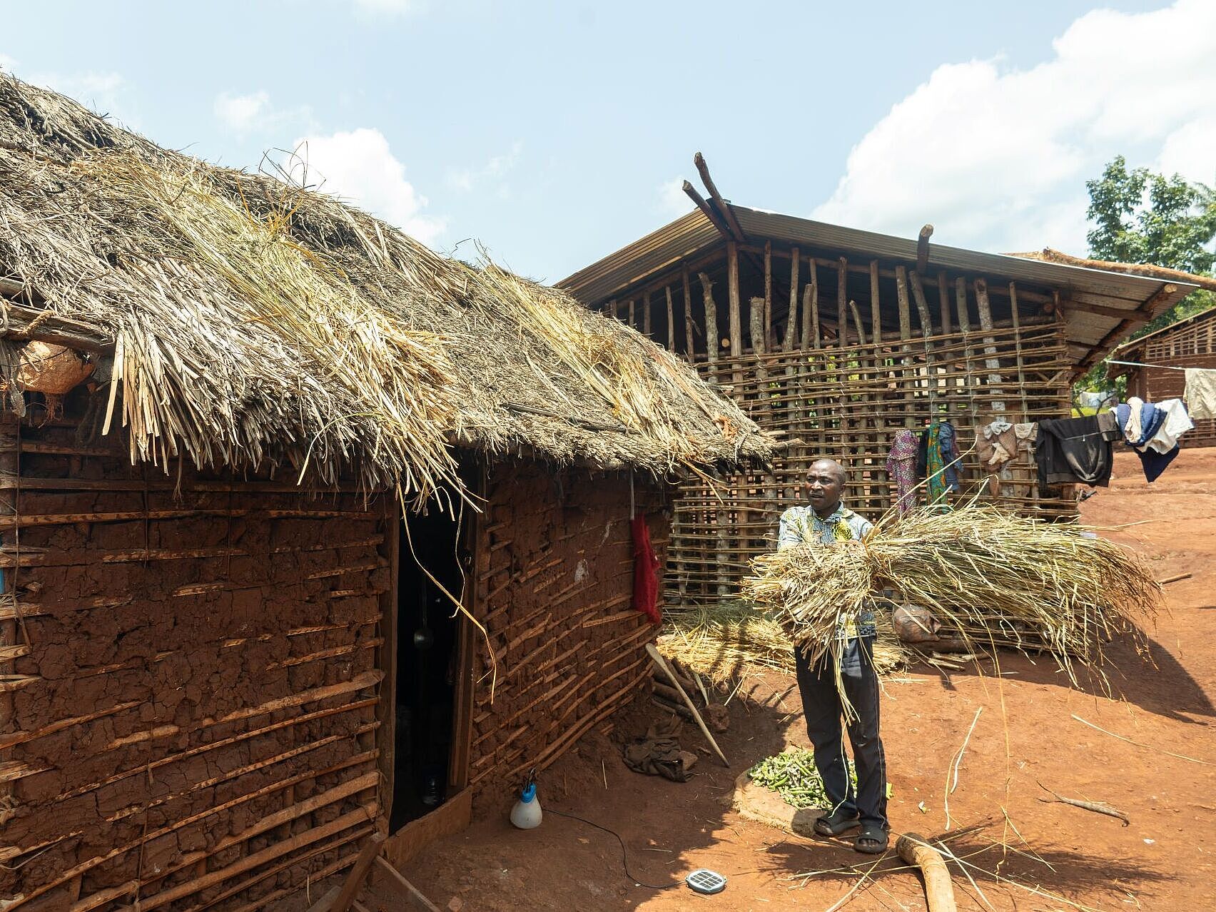 Ein mann steht vor einer selbst gebauten Hütte und verstärkt das Dach mit Stroh