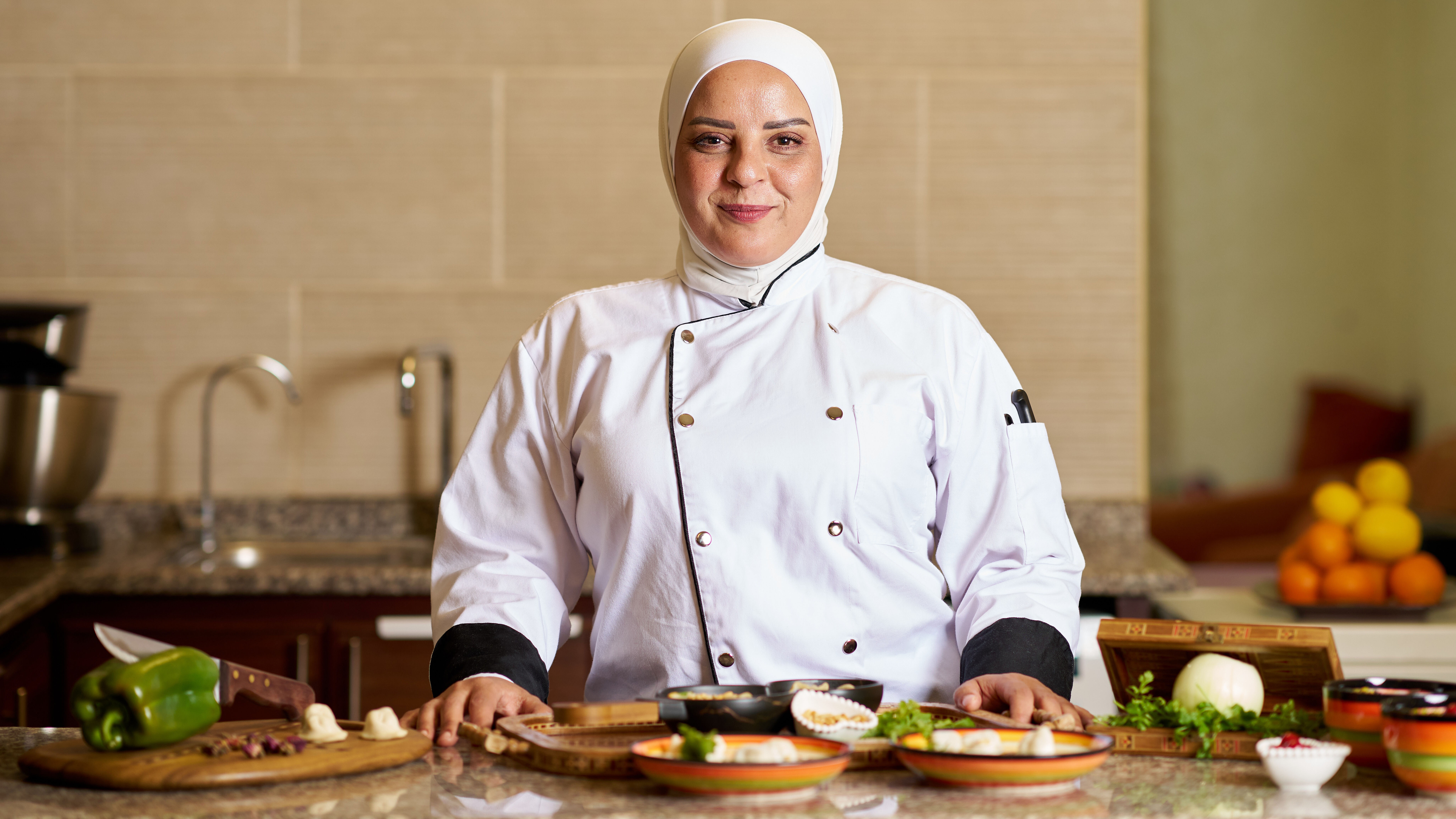 Mouna steht lächelnd in Kochschürze vor ihren selbst gekochten Köstlichkeiten