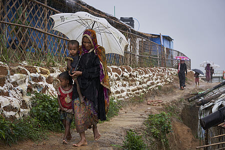 Eine Frau und zwei Kinder gehen mit einem Regenschirm durch das Flüchtlingscamp Kutupalong.
