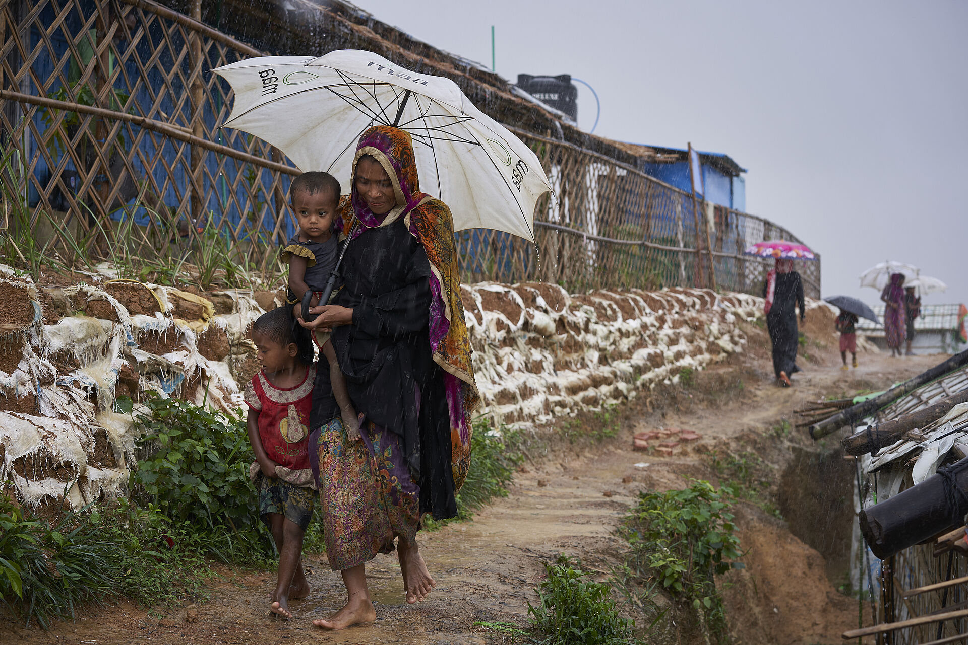  Eine Frau und zwei Kinder gehen mit einem Regenschirm durch das Flüchtlingscamp Kutupalong. © UNHCR/David Azia  Rohingya-Flüchtlinge bahnen sich während eines heftigen Monsunregens im Flüchtlingscamp Kutupalong ihren Weg