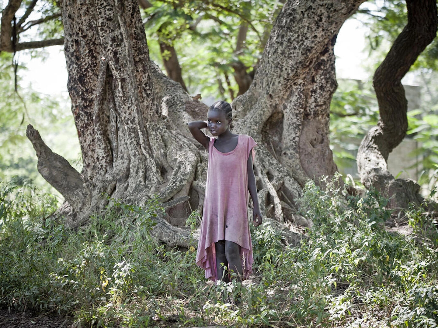 Afrikanisches Mädchen steht in zerschlissenem roten Kleid in einem Wald.