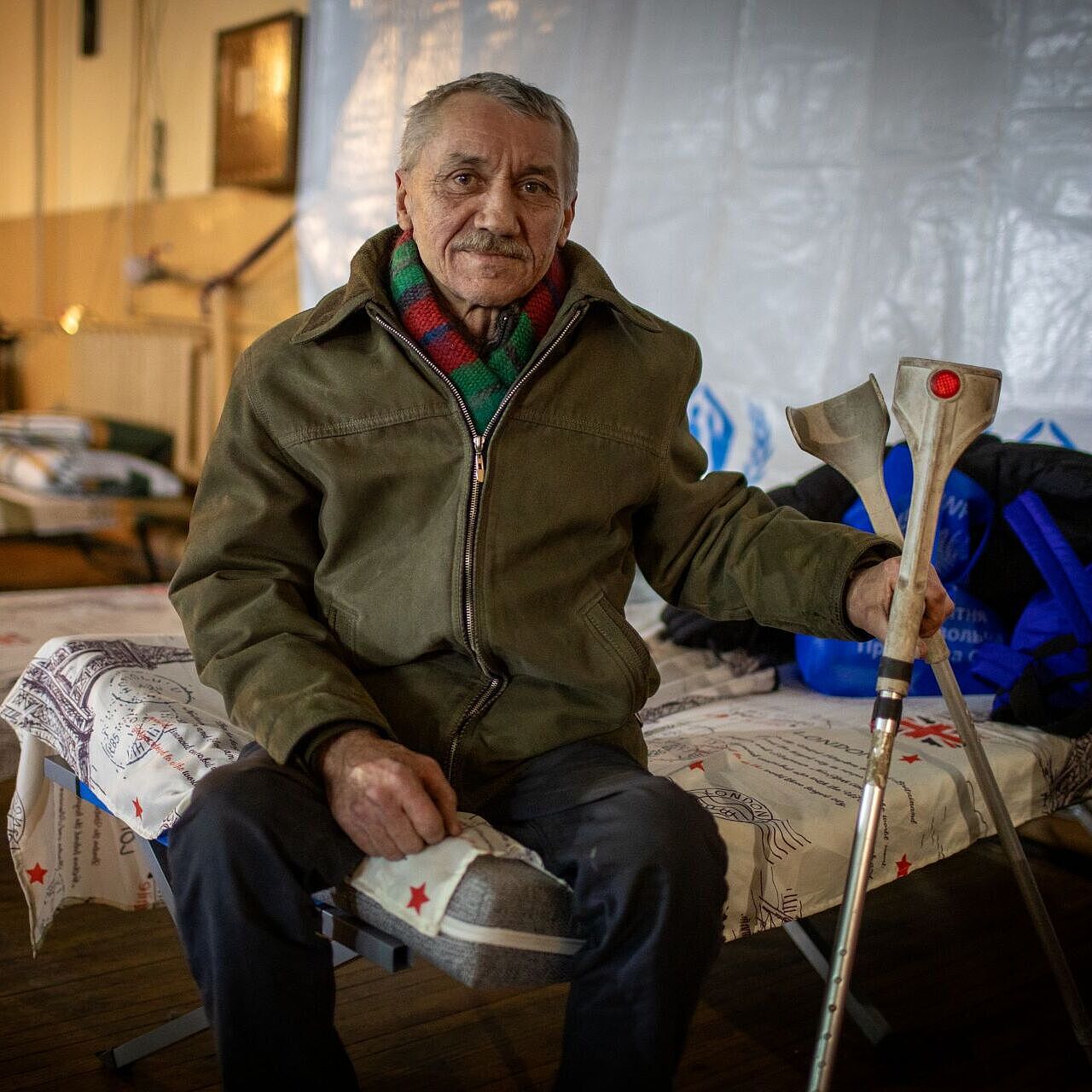 Ein alter Mann sitzt auf seiner Pritsche, Krücken in der Hand.