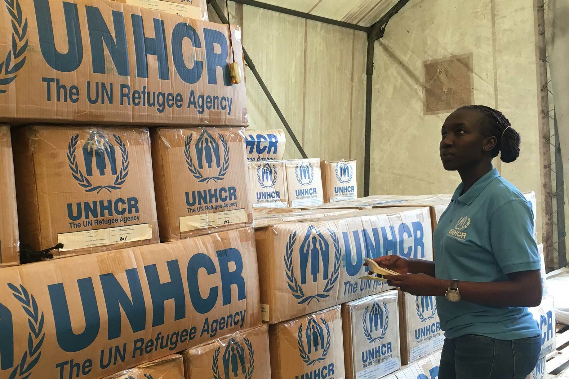 Robina in einem Lage mit Hilfsgütern des UNHCR. Sie hat eine Liste in der Hand und scheint die Kistenanzahl zu kontrolieren. 