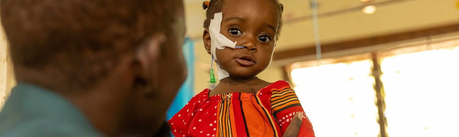 Ein kleines Mädchen im bunten Kleid wird von seinem Vater gehalten und hat medizinische Schläuche am Gesicht